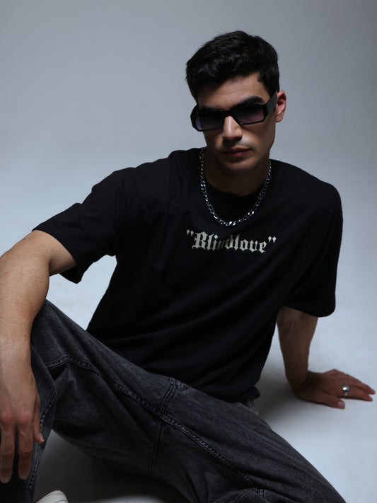 Male model seated in a black Joan Alvi BlindLove oversized T-shirt, showing the small Gothic chest logo and relaxed fit