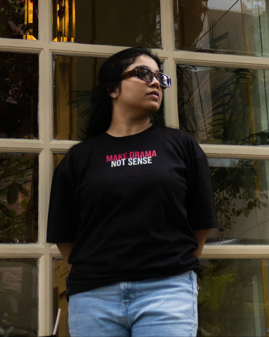 A woman stands outdoors in a relaxed fit pose, wearing a premium black cotton t-shirt featuring an oversized fit. The t-shirt showcases high quality print across the chest that reads ‘MAKE DRAMA NOT SENSE’ in bold red and white lettering. Crafted from heavy 240 gsm french terry cotton, the oversized t-shirt is paired with light blue jeans.