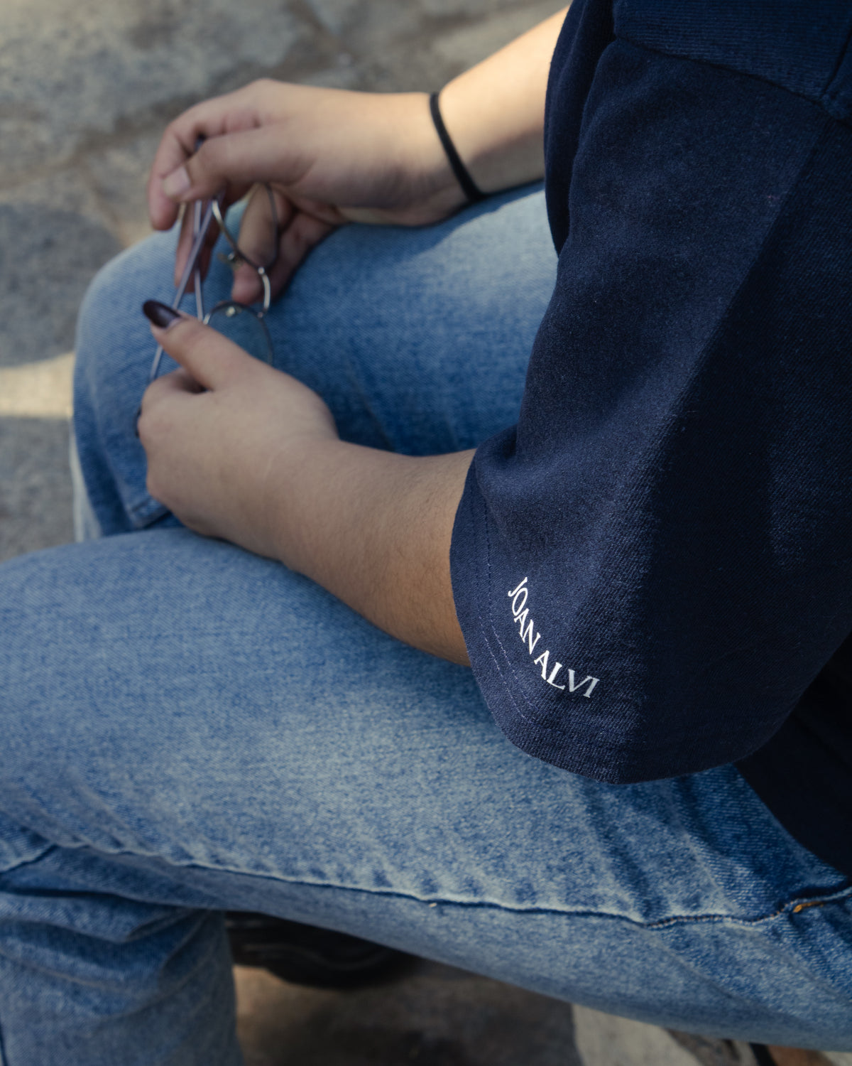 A close-up shot focusing on the sleeve of a premium navy blue oversized t-shirt, highlighting its high-quality French Terry cotton texture. The sleeve features the minimalist white text branding "JOAN ALVI" embroidered near the hem. A person is sitting in a relaxed fit pose wearing light blue denim jeans, with their hands resting in their lap holding sunglasses.