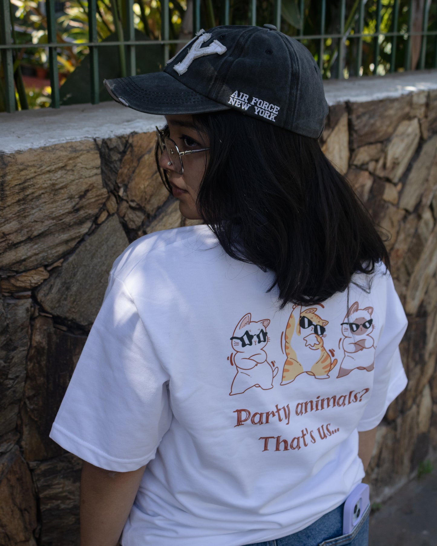 Rear view of a person wearing a white, heavy French Terry cotton oversized t-shirt featuring a relaxed fit and a high-quality graphic print on the back showing three cartoon cats wearing sunglasses above the text "Party animals? That's us...".