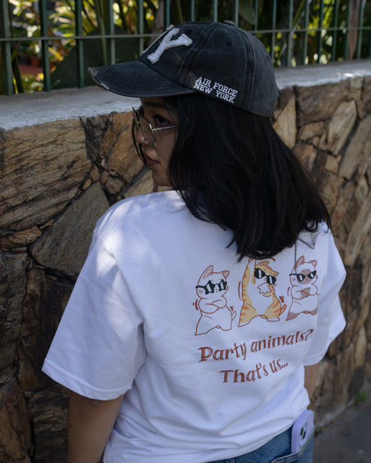 Rear view of a person wearing a white, heavy French Terry cotton oversized t-shirt featuring a relaxed fit and a high-quality graphic print on the back showing three cartoon cats wearing sunglasses above the text "Party animals? That's us...".