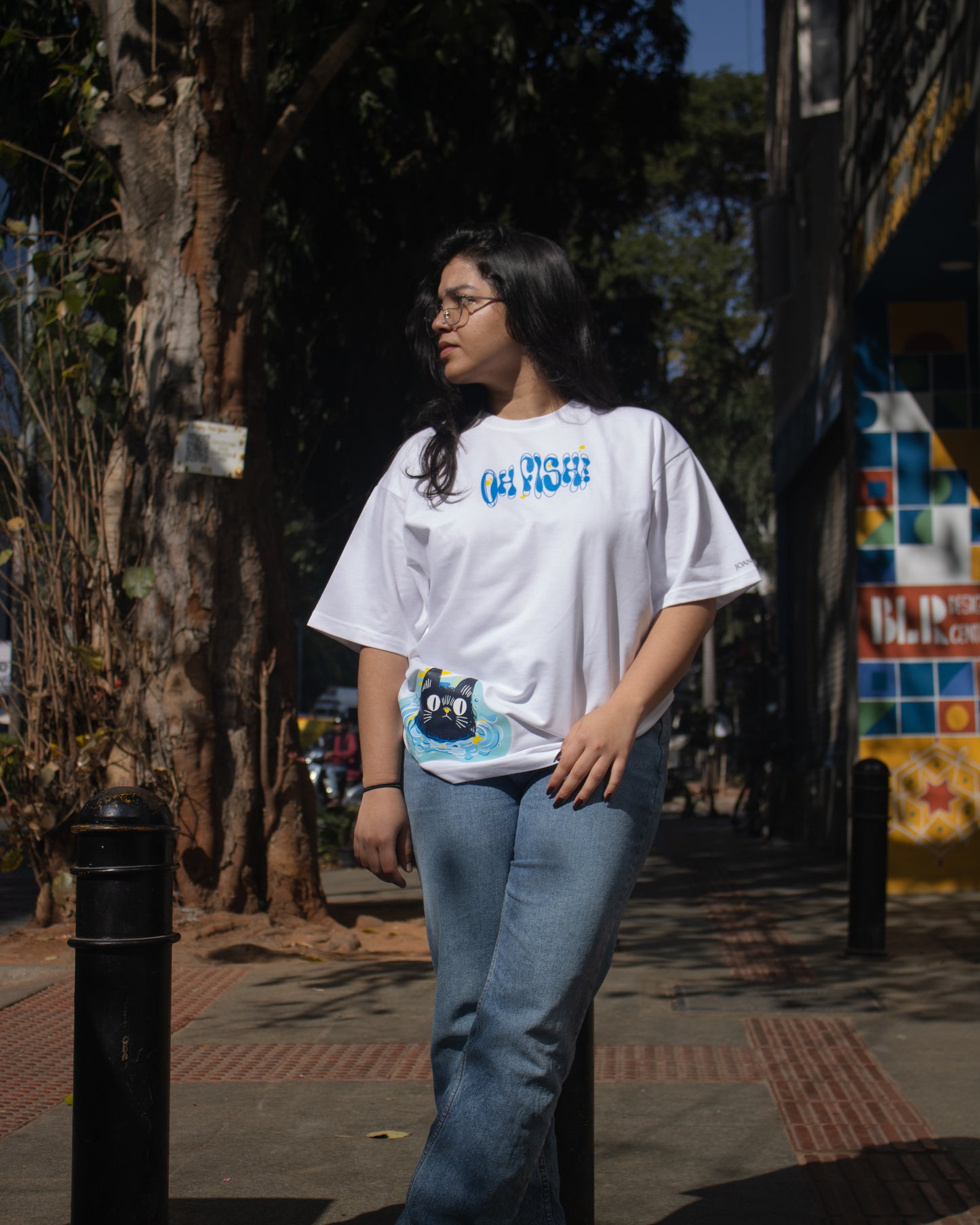 A full-length outdoor shot of a person wearing a white premium French Terry cotton oversized t-shirt with a relaxed fit and blue jeans, standing on a sunny sidewalk. The tee displays a high-quality graphic print with blue text that reads "OH FISH!" across the chest and a cartoon cat illustration near the lower hem.