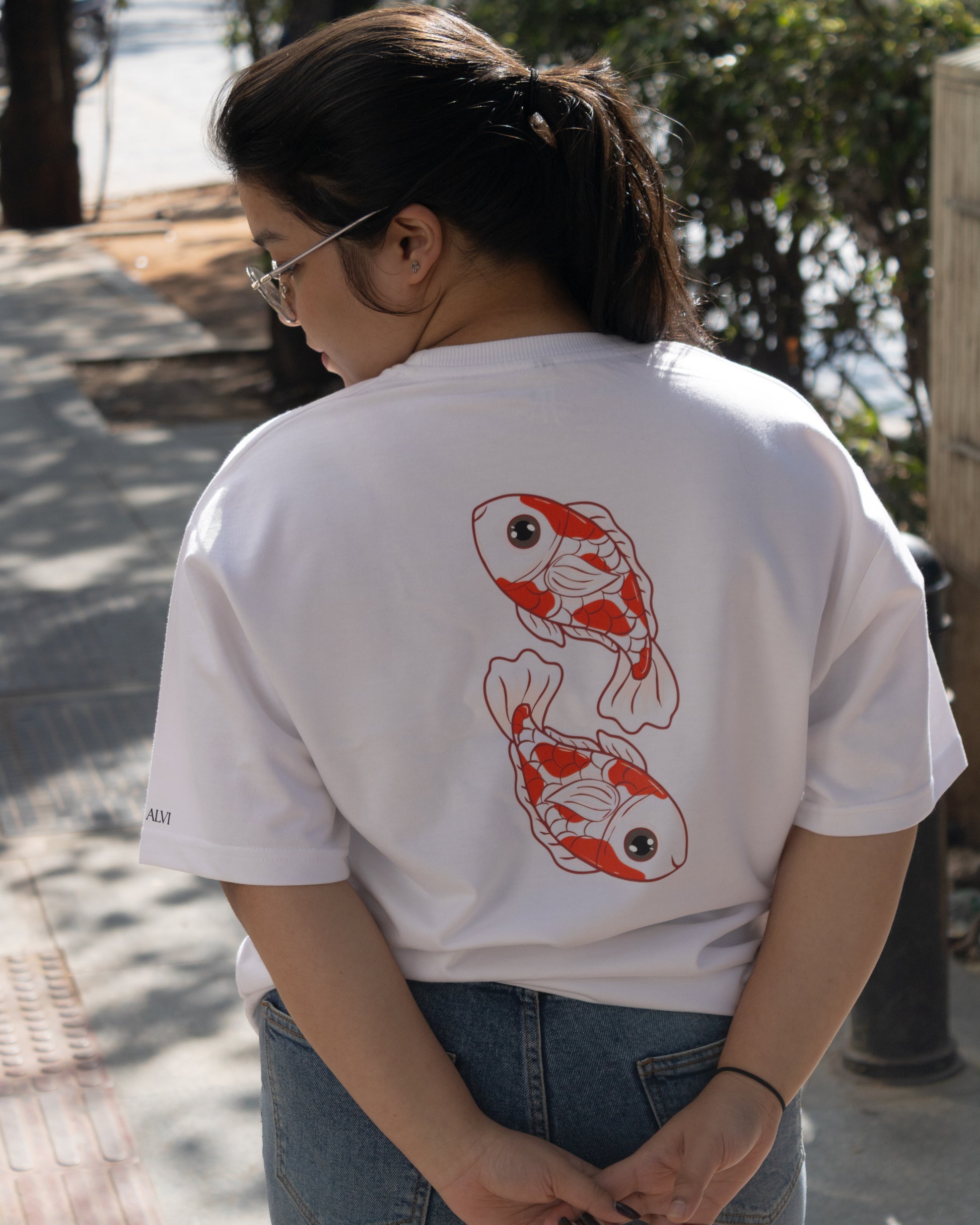 Rear view of a person standing outdoors wearing a white premium French Terry cotton oversized t-shirt with a relaxed fit and blue jeans. The back of the tee highlights a high-quality graphic print depicting two stylized orange and white koi fish swimming in a circular formation.