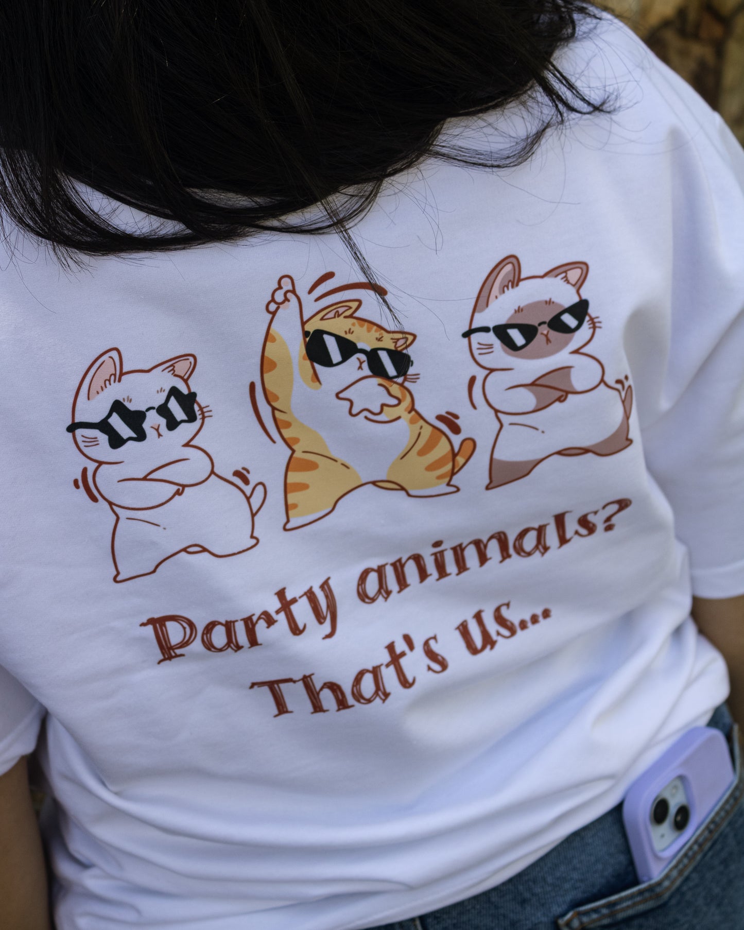 Close-up rear view of a person wearing a white, heavy French Terry cotton oversized t-shirt with a relaxed fit, highlighting a high-quality graphic print on the back featuring three cartoon cats wearing black sunglasses dancing above the text "Party animals? That's us...".