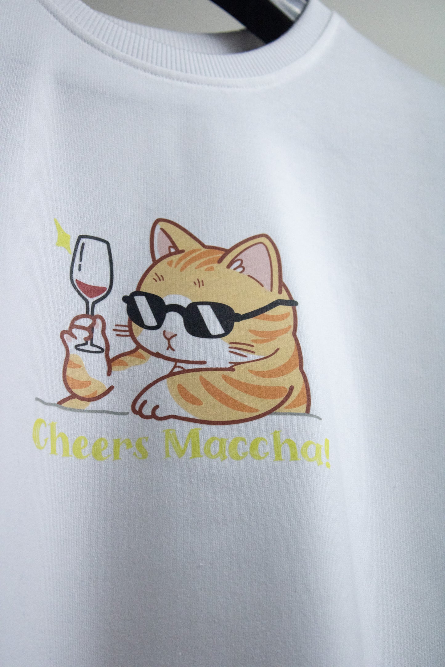 Close-up view of a white premium French Terry cotton oversized t-shirt with a relaxed fit, showcasing a high-quality graphic print on the front of a cool orange tabby cat wearing black sunglasses and raising a wine glass above the yellow text "Cheers Maccha!".