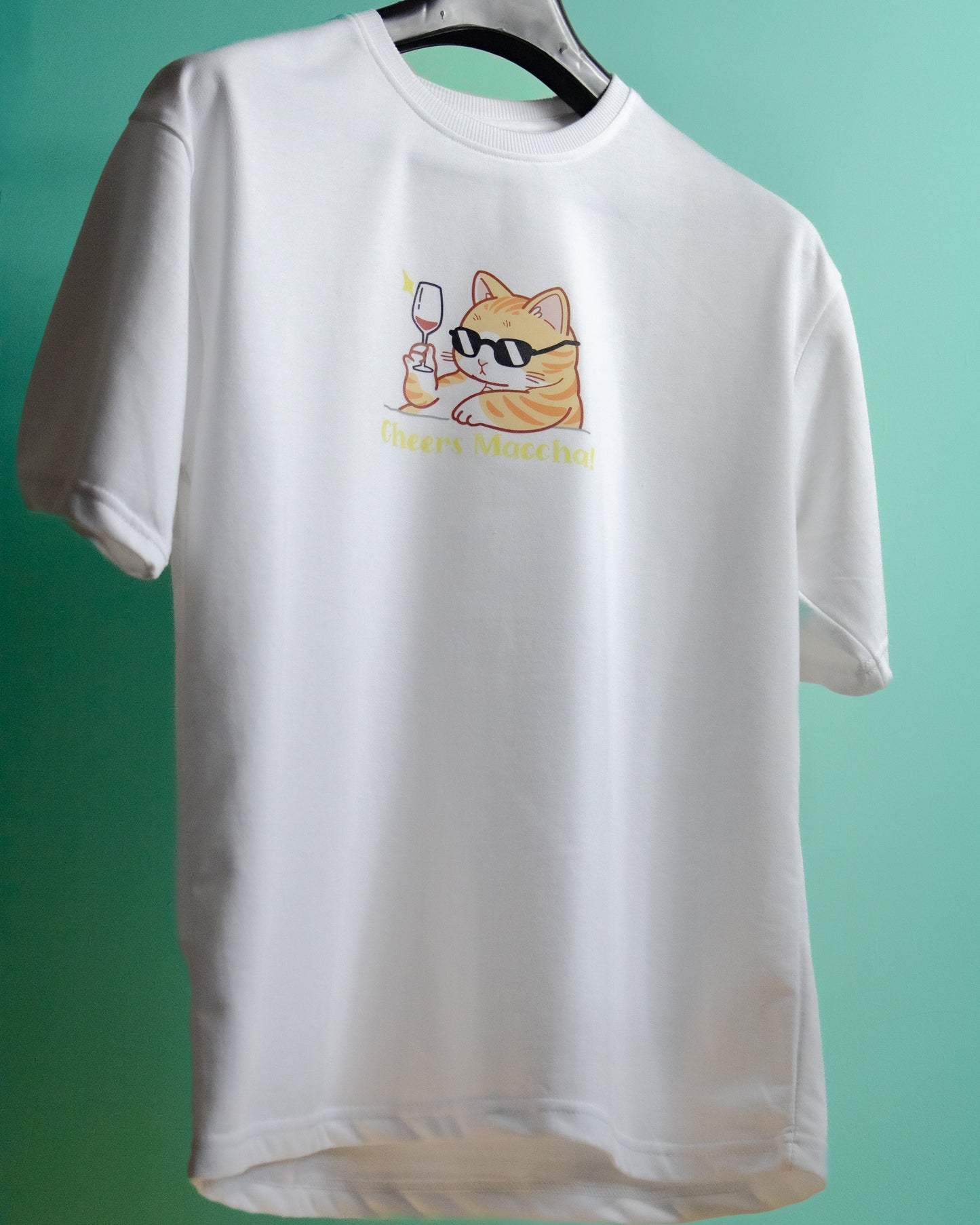 A white premium French Terry cotton oversized t-shirt with a relaxed fit hanging on a hanger against a bright green background, featuring a high-quality graphic print on the front of an orange tabby cat wearing black sunglasses and holding a wine glass above the text "Cheers Maccha!".