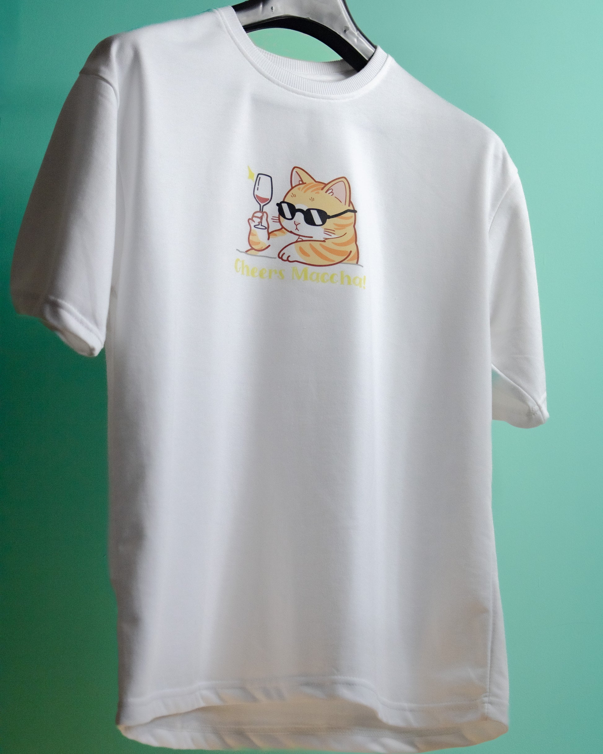 A white premium French Terry cotton oversized t-shirt with a relaxed fit hanging on a hanger against a bright green background, featuring a high-quality graphic print on the front of an orange tabby cat wearing black sunglasses and holding a wine glass above the text "Cheers Maccha!".
