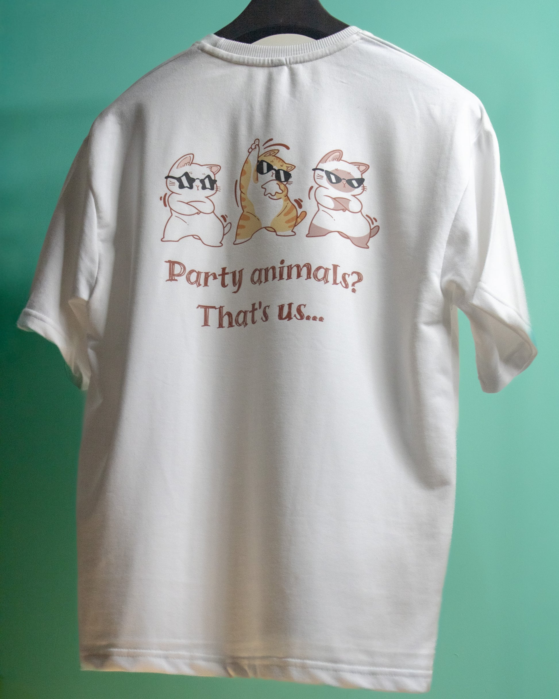 A full rear view of a white premium French Terry cotton oversized t-shirt with a relaxed fit hanging on a clothes hanger against a bright green background, showing a high-quality graphic print of three dancing cartoon cats wearing sunglasses above the text "Party animals? That's us...".