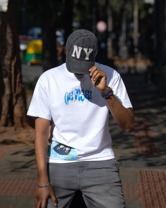 Front view of a person standing outdoors wearing a white premium French Terry cotton oversized t-shirt with a relaxed fit and grey pants, accessorized with a dark grey "NY" baseball cap. The tee features a high-quality graphic print with wavy blue text reading "OH FISH!" across the chest and a small cartoon cat illustration near the bottom hem.