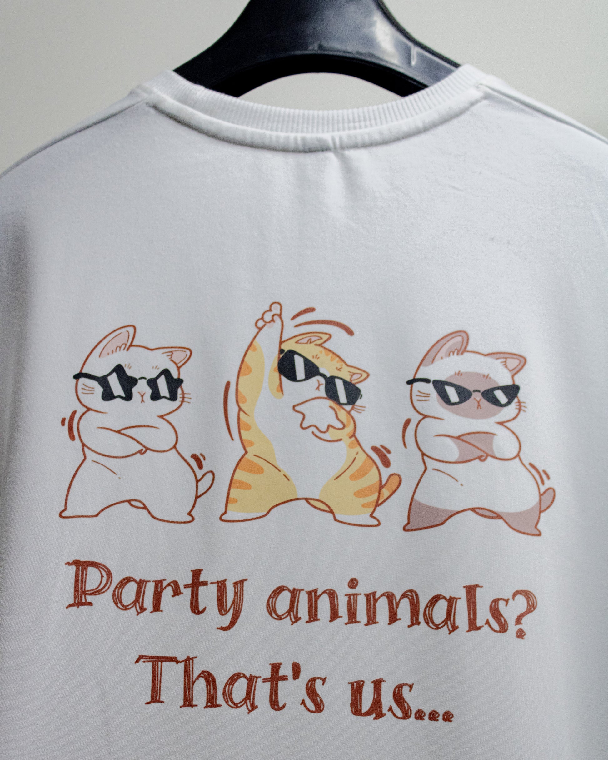 Close-up rear view of a white premium French Terry cotton oversized t-shirt hanging on a clothes hanger, displaying a high-quality graphic print on the back featuring three cartoon cats wearing black sunglasses dancing above the text "Party animals? That's us...".